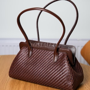 Op de afbeelding: Bruine leren handtas met een geweven textuur. De tas heeft twee gebogen handvatten en een rechthoekig frame. Het woord "NOSTALGIA" is zichtbaar op het frame. De tas staat op een houten oppervlak.