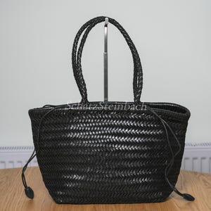 Può includere: Una borsa a mano nera intrecciata con una base strutturata e due manici intrecciati. La borsa presenta una chiusura a coulisse con sottili cordini neri che terminano con piccoli fermi neri su ciascun lato. Il testo SchatzSteinbach è stampato in bianco.