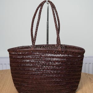 Puede incluir: Un bolso de mano trenzado de color marrón oscuro con un patrón de espiga en todo el cuerpo y dos asas trenzadas. El texto SchatzSteinbach está impreso en un color claro en la parte superior central del bolso, que tiene un diseño abierto.