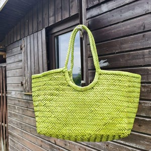 Pode incluir: Uma bolsa tote verde-limão com um padrão de espinha de peixe tecido em todo o corpo. A bolsa inclui duas alças trançadas presas à abertura superior, criando um design estruturado de abertura superior com uma textura consistente.