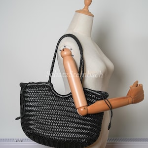 May include: Black woven leather tote bag with braided handles. The bag has a rounded shape and a woven pattern. The bag is displayed on a mannequin. The bag is a dark colour.