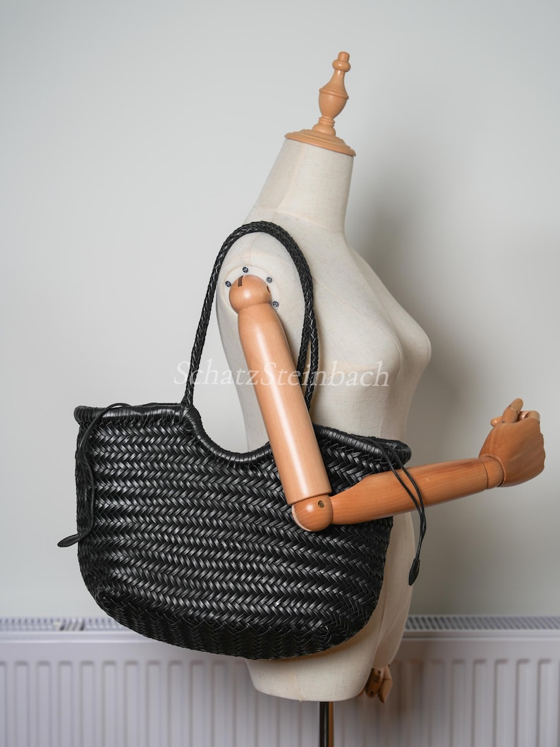 May include: Black woven leather tote bag with braided handles. The bag has a rounded shape and a woven pattern. The bag is displayed on a mannequin. The bag is a dark colour.