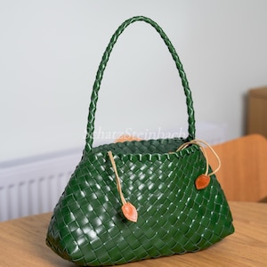 May include: A green woven handbag with a braided handle. The bag has a trapezoid shape and is made of a tightly woven material. Tan leather accents are attached to the bag with drawstrings. The bag is sitting on a wooden table.