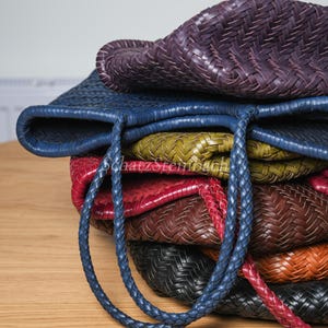 May include: A pile of woven leather tote bags in shades of purple, blue, green, red, brown, black, and orange. The bags feature braided handles and a basket-weave texture. The bags are stacked on a wooden surface.