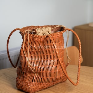 Può includere: Una borsa a secchiello intrecciata marrone con esterno a griglia, due tracolle intrecciate e una sacca interna beige con coulisse. Il testo SchatzSteinbach è stampato al centro della borsa con un carattere chiaro e semi-trasparente.