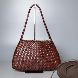 May include: A brown leather handbag with a woven design. The bag has a braided handle and a matching small pouch. The bag is displayed on a silver stand against a neutral background.