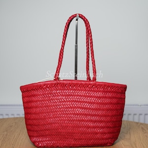 May include: A vibrant red woven tote bag featuring a distinct herringbone pattern throughout the body. The bag includes two long, braided red handles attached to the top rim. The text SchatzSteinbach is visible in white lettering across the center.