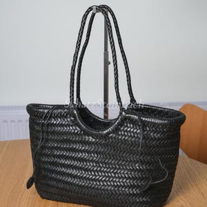 May include: Black woven leather tote bag with braided handles. The bag has a rectangular shape with a woven pattern and a drawstring closure. The handles are long enough to be worn over the shoulder. The bag is sitting on a wooden table.