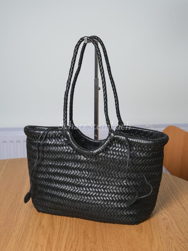 May include: Black woven leather tote bag with braided handles. The bag has a rectangular shape with a woven pattern and a drawstring closure. The handles are long enough to be worn over the shoulder. The bag is sitting on a wooden table.