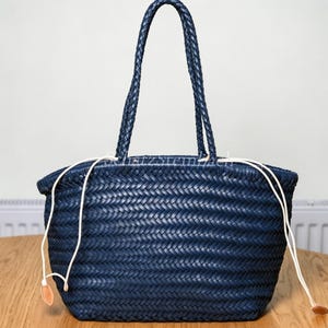 Op de afbeelding: Een marineblauwe gevlochten tas met een geweven textuur, twee bijpassende gevlochten schouderbanden en een witte trekkoordsluiting met kleine lichtbruine lederachtige lipjes aan de uiteinden. De tas heeft een brede bodem en een open ontwerp.
