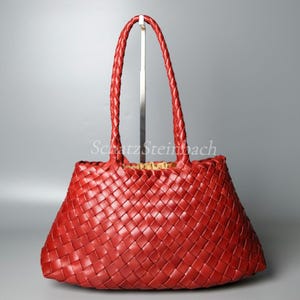 Può includere: Una borsa tote rossa brillante con un motivo a intreccio su tutto l'esterno. La borsa include due manici intrecciati e una fodera interna color marrone chiaro visibile all'apertura superiore. Il testo SchatzSteinbach è centrato.