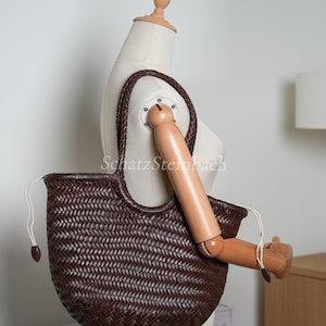 May include: A dark brown woven tote bag with a braided handle. The bag has a rounded shape and a drawstring closure. The bag is displayed on a mannequin.