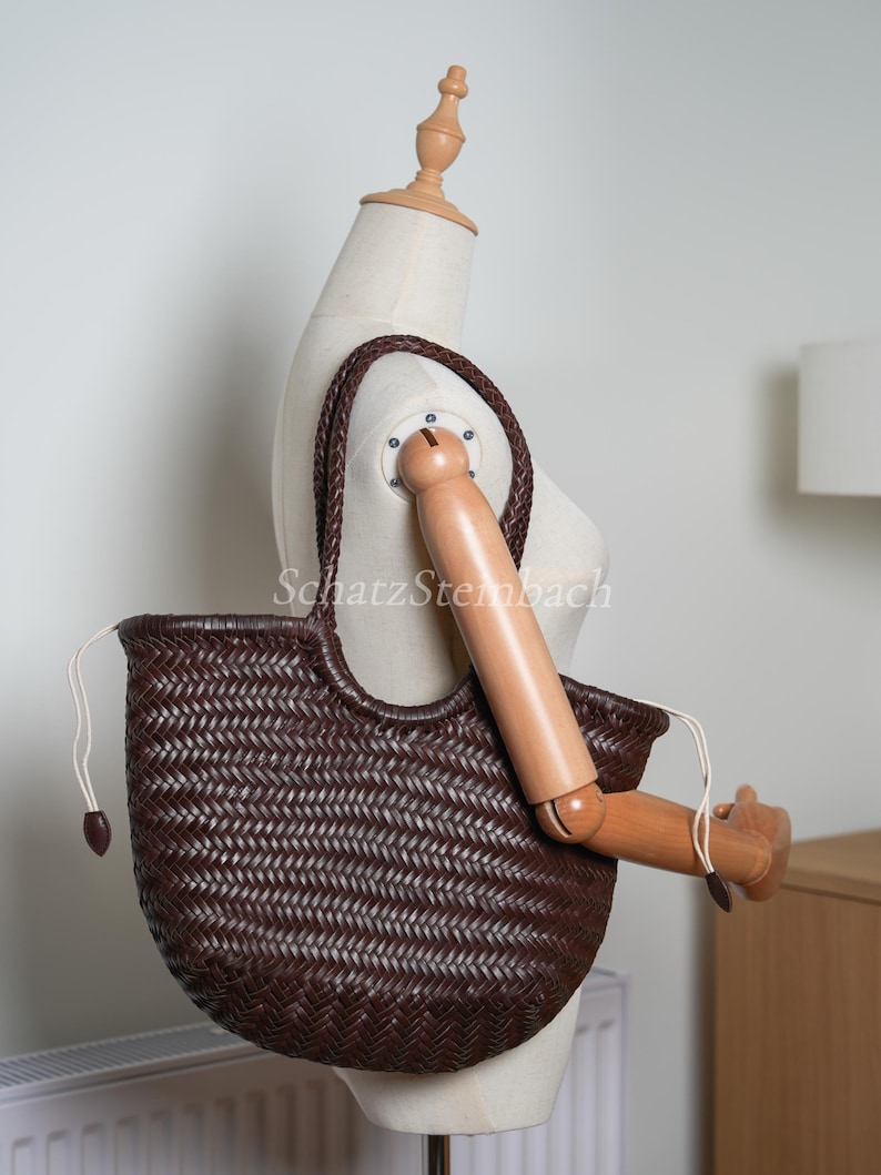 May include: A dark brown woven tote bag with a braided handle. The bag has a rounded shape and a drawstring closure. The bag is displayed on a mannequin.