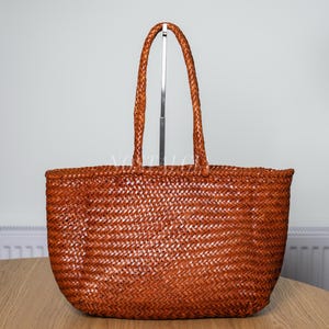 May include: A medium brown tote bag featuring a woven construction throughout the body and two matching braided handles. The bag has a rectangular shape with rounded bottom corners and a consistent, tight weave pattern across the entire surface.