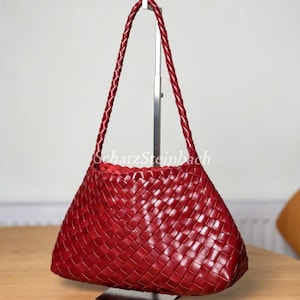 May include: A deep red handbag featuring a woven leather exterior pattern and a single braided shoulder strap. The bag has a structured, trapezoidal shape with a smooth finish and the text SchatzSteinbach printed in white across the center.