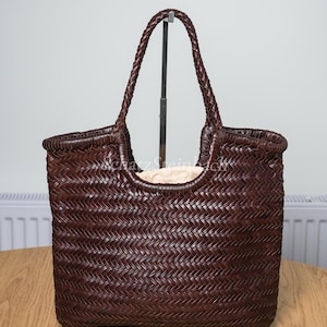 Può includere: Una borsa tote in pelle intrecciata marrone scuro con motivo a spina di pesce, due manici intrecciati e una fodera interna in tessuto beige chiaro. Il testo SchatzSteinbach è visibile in lettere bianche al centro della borsa.