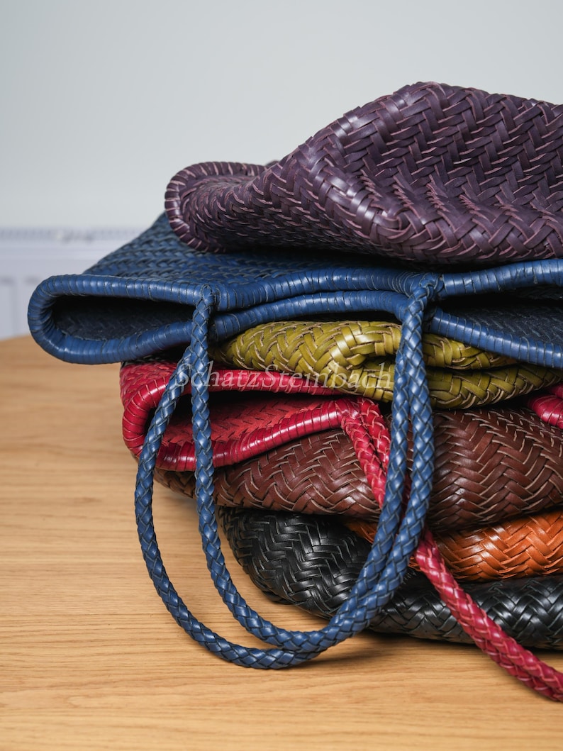 May include: A collection of woven leather tote bags in shades of purple, blue, green, red, brown, black, and orange. The bags feature braided handles and a basket-weave texture. They are stacked on a wooden surface.