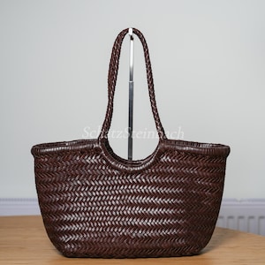 May include: A dark brown woven leather tote bag with braided handles. The bag has a rounded shape and a textured surface. The handles are attached to the top of the bag with a metal support.