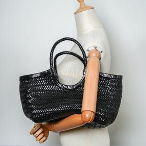 May include: A black woven tote bag with braided handles. The bag has a rectangular shape and a textured surface. The bag is held by a wooden mannequin arm. The bag is a stylish accessory.