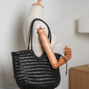 May include: Black woven leather tote bag with braided handles. The bag has a unique basket weave design and a spacious interior. The bag is displayed on a mannequin.