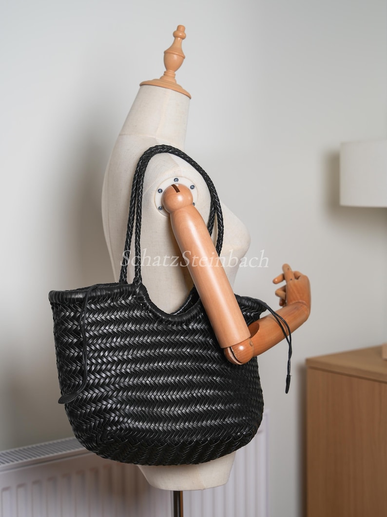 May include: Black woven leather tote bag with braided handles. The bag has a unique basket weave design and a spacious interior. The bag is displayed on a mannequin.