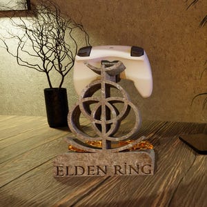 May include: A grey and gold Elden Ring video game controller stand. The stand has a decorative design and holds a white controller. The base of the stand has the words "ELDEN RING" engraved on it.