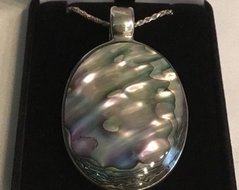 May include: Oval pendant necklace with a shimmering, iridescent abalone shell set in a silver-toned metal frame. The pendant is displayed in a black velvet jewelry box, showcasing its natural colors of green, purple, and silver.