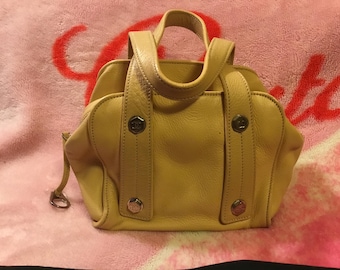 May include: A light yellow leather handbag with two handles and silver-tone hardware. The bag has a structured shape with a top handle and two vertical straps with decorative buttons. The bag is sitting on a pink surface.