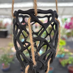 May include: A black, open-weave hanging planter suspended by natural fiber rope. The planter has a geometric design and is designed to hold a small potted plant. The background is blurred, suggesting an outdoor setting.