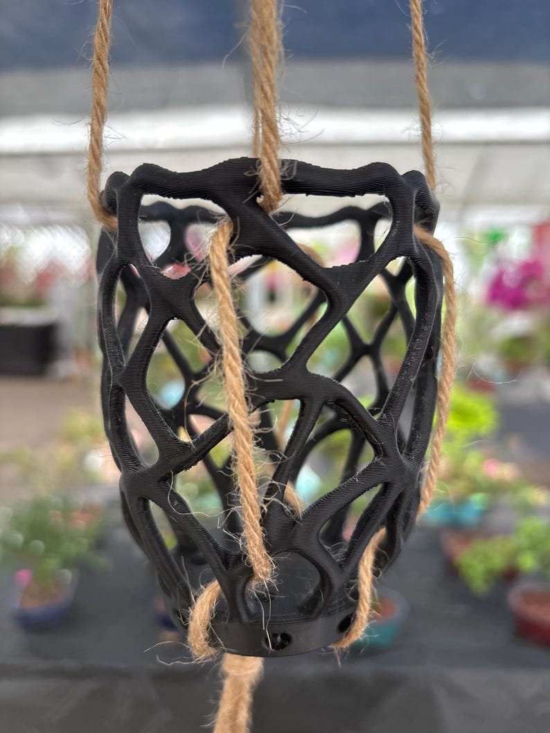 May include: A black, open-weave hanging planter suspended by natural fiber rope. The planter has a geometric design and is designed to hold a small potted plant. The background is blurred, suggesting an outdoor setting.