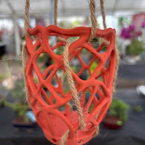 May include: A vibrant orange hanging planter with a lattice design. The planter is suspended by three natural-colored rope cords. The openwork design allows for air circulation and drainage. Ideal for displaying small plants or flowers. The planter's dimensions are approximately 6 inches in diameter and 5 inches in height.