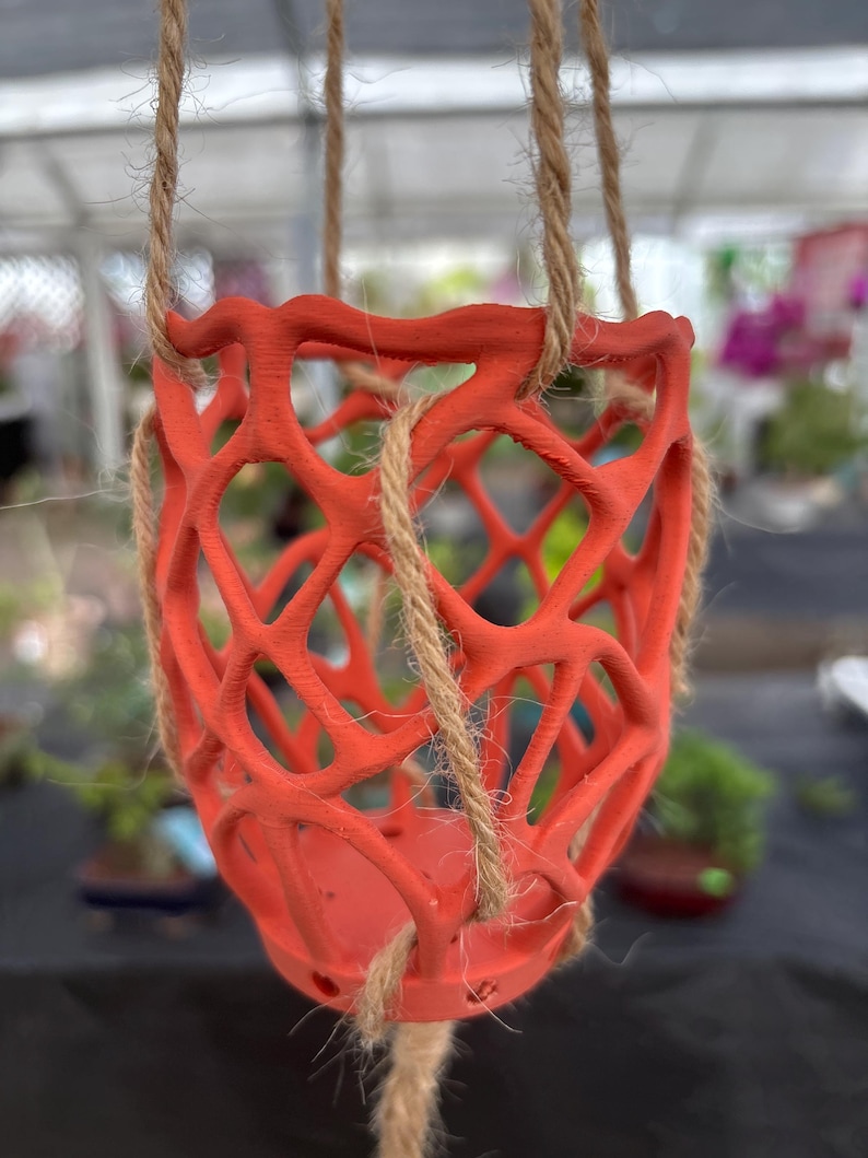 May include: A vibrant orange hanging planter with a lattice design. The planter is suspended by three natural-colored rope cords. The openwork design allows for air circulation and drainage. Ideal for displaying small plants or flowers. The planter's dimensions are approximately 6 inches in diameter and 5 inches in height.