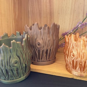 May include: Three decorative tree-trunk-style planters in shades of green, brown, and orange. The planters have a textured wood grain appearance with vertical slits and a jagged top edge. They are displayed on a wooden shelf.