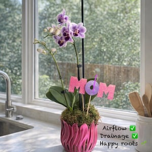 May include: A potted orchid with purple and white flowers. The pink pot has a unique, wavy design. A glittery pink sign that says "MOM" is inserted into the pot. The text "Airflow, Drainage, Happy roots" is also visible.