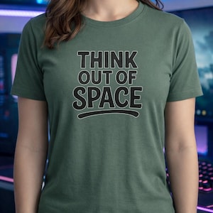 Könnte beinhalten: Ein kurzärmeliges, dunkelgrünes T-Shirt mit dem Text "THINK OUT OF SPACE" in fetten, schwarzen Buchstaben. Der Text ist zentriert und mit einer schwarzen Linie unterstrichen. Das T-Shirt ist aus weichem Material.