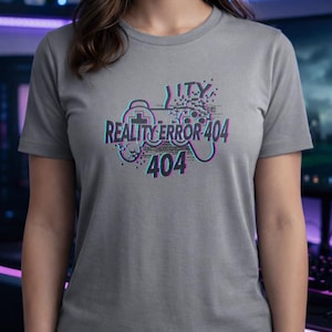 Può includere: Una maglietta grigia con un design a effetto glitch. Il design include un controller di gioco e il testo "REALITY ERROR 404" in verde acqua, rosa e nero. La maglietta è a maniche corte.