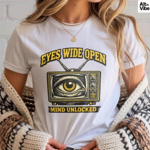 May include: White t-shirt featuring a yellow and black graphic of a vintage television with an eye in the screen. The text "EYES WIDE OPEN" is above the television, and "MIND UNLOCKED" is below.