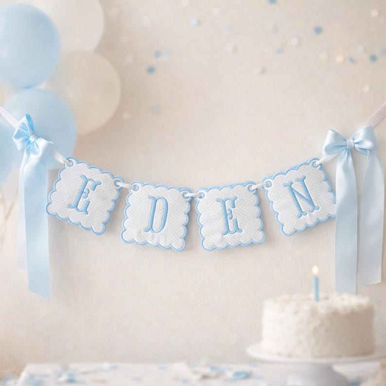 May include: A banner spelling "EDEN" in blue embroidered letters hangs against a white wall, with blue and white balloons and a small cake with a lit candle. The banner has a scalloped edge and is tied with blue ribbons.