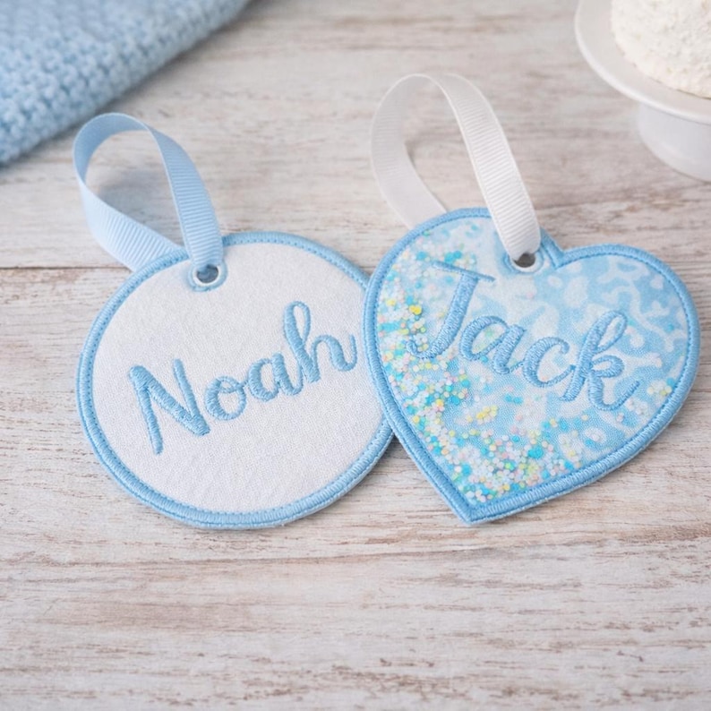 May include: Two personalized gift tags. One is round with the name "Noah" embroidered in light blue. The other is heart-shaped with "Jack" embroidered in light blue, with a blue and white pattern and white ribbon.