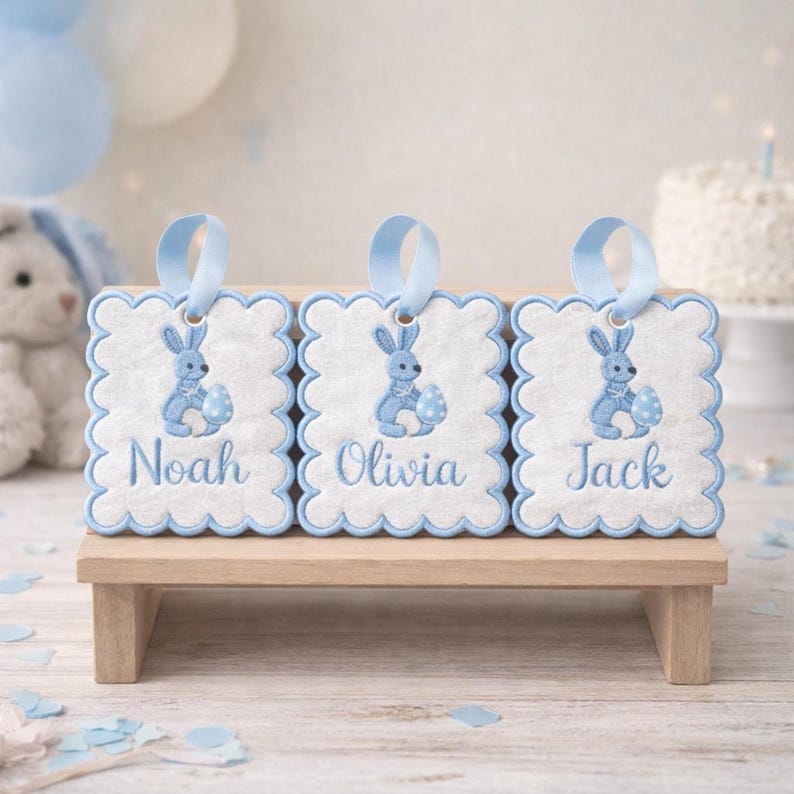 May include: Three embroidered tags with blue scalloped edges, each featuring a blue bunny holding a decorated egg. The names "Noah", "Olivia", and "Jack" are embroidered below the bunny. Each tag has a blue ribbon loop for hanging. The tags are displayed on a small wooden stand.