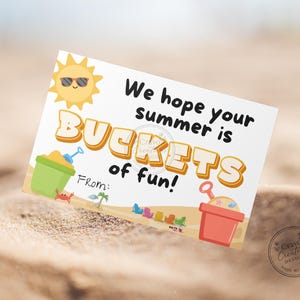 Puede incluir: Una tarjeta blanca con el texto "We hope your summer is BUCKETS of fun!" presenta un sol de dibujos animados con gafas de sol, cubos de arena y sillas de playa. La tarjeta está sobre una superficie arenosa.