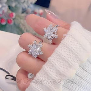 Könnte beinhalten: Zwei silberne Ringe mit Schneeflocken-Designs, jeweils mit klaren, funkelnden Kristallen besetzt. Die Ringe werden an Fingern vor einem weichen, weißen Hintergrund gezeigt. Der Text "SILVERMARSTER" ist unten sichtbar.