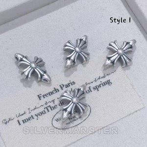 May include: Four silver-colored cross-shaped charms. The charms have a detailed, ornate design with a central circular element and four outward-facing arms. The text "Style I" is visible in the upper right corner. A card with text is in the image.