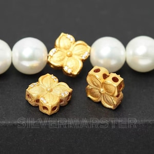 May include: Close-up of gold-colored flower-shaped beads with small clear stones. The beads are next to large white pearls. The text "SILVERMARSTER" is at the bottom of the image.