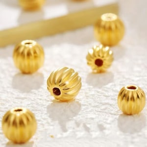 May include: Close-up of several gold-colored beads with a ribbed design. The beads are scattered on a white surface. The beads have a small hole in the center, and the light reflects off the shiny surface.