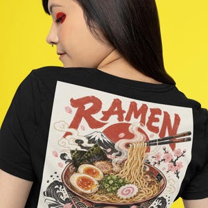 Könnte beinhalten: Ein schwarzes T-Shirt mit einem großen Grafikdruck auf der Rückseite. Das Design zeigt eine Schüssel Ramen mit Nudeln, weich gekochten Eiern und Frühlingszwiebeln, garniert mit Stäbchen. Der Text RAMEN steht über einem Berg und einer roten Sonne.
