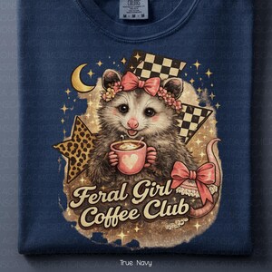 May include: Navy blue sweatshirt featuring a possum with a floral crown and bow, holding a coffee cup with a heart. The design includes a crescent moon, stars, and the text "Feral Girl Coffee Club".