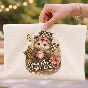 May include: A cream-colored pouch featuring a possum illustration with a floral crown, holding a coffee cup. The text "Feral Girl Coffee Club" is below the possum. The design includes a checkered flag and a crescent moon.