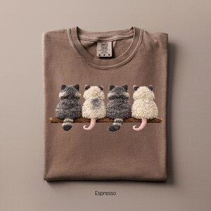 May include: Espresso-colored t-shirt with a crew neck. The shirt features an embroidered design of four possums sitting on a branch. The possums are in shades of gray and white, with pink tails. The shirt has a Comfort Colors tag.