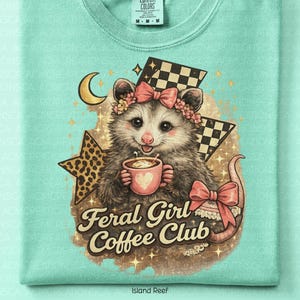 May include: A light turquoise t-shirt featuring a possum illustration holding a coffee cup. The possum wears a floral crown and a pink bow. The text "Feral Girl Coffee Club" is below the possum. A crescent moon, stars, and checkered flags complete the design.